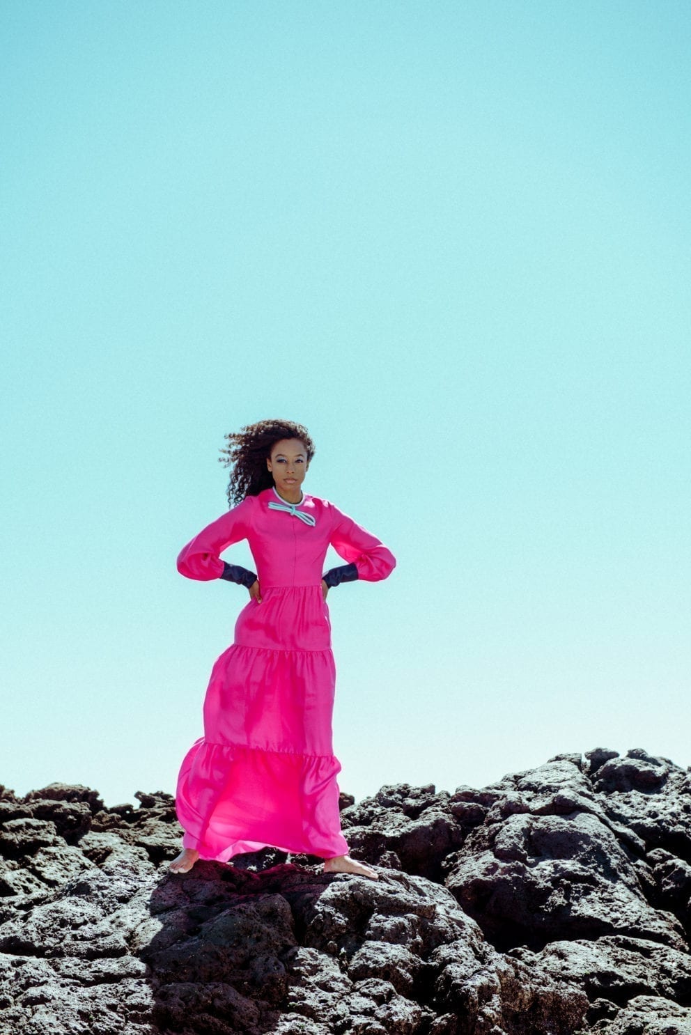 Corinne Bailey Rae in pink dress standing against blue sky on rocks