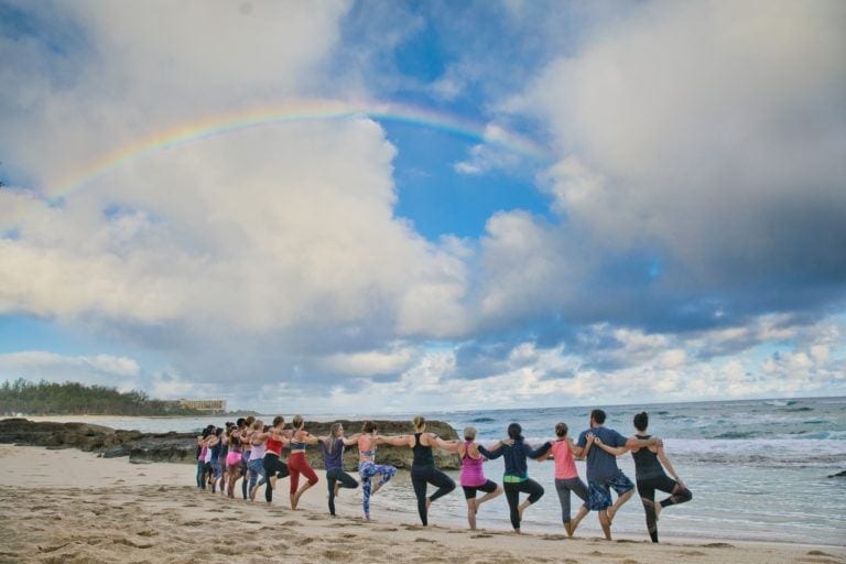 people doing yoga tree pose under a rainbow on beach